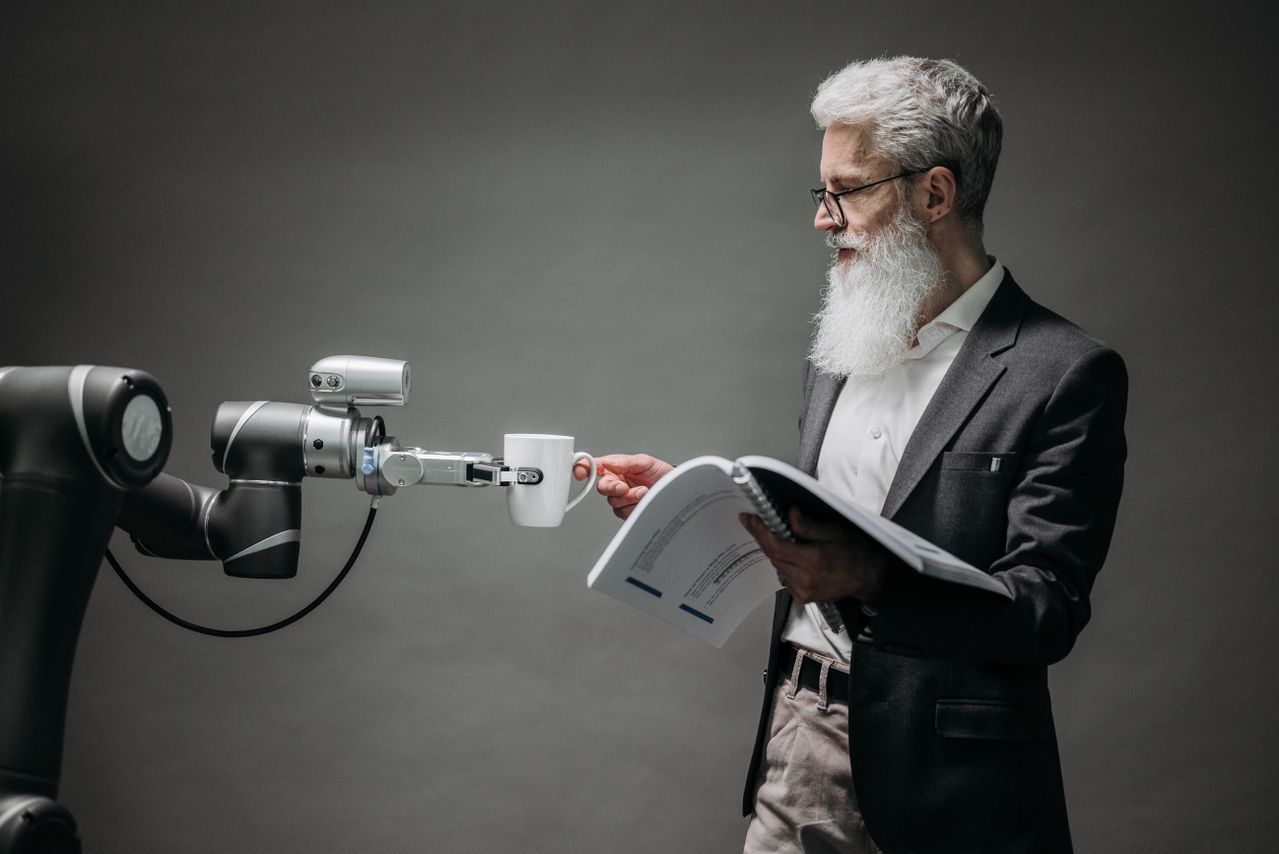 Een robotarm reikt een kop koffie aan aan een man met een witte baard en bril, die ondertussen een rapport doorneemt