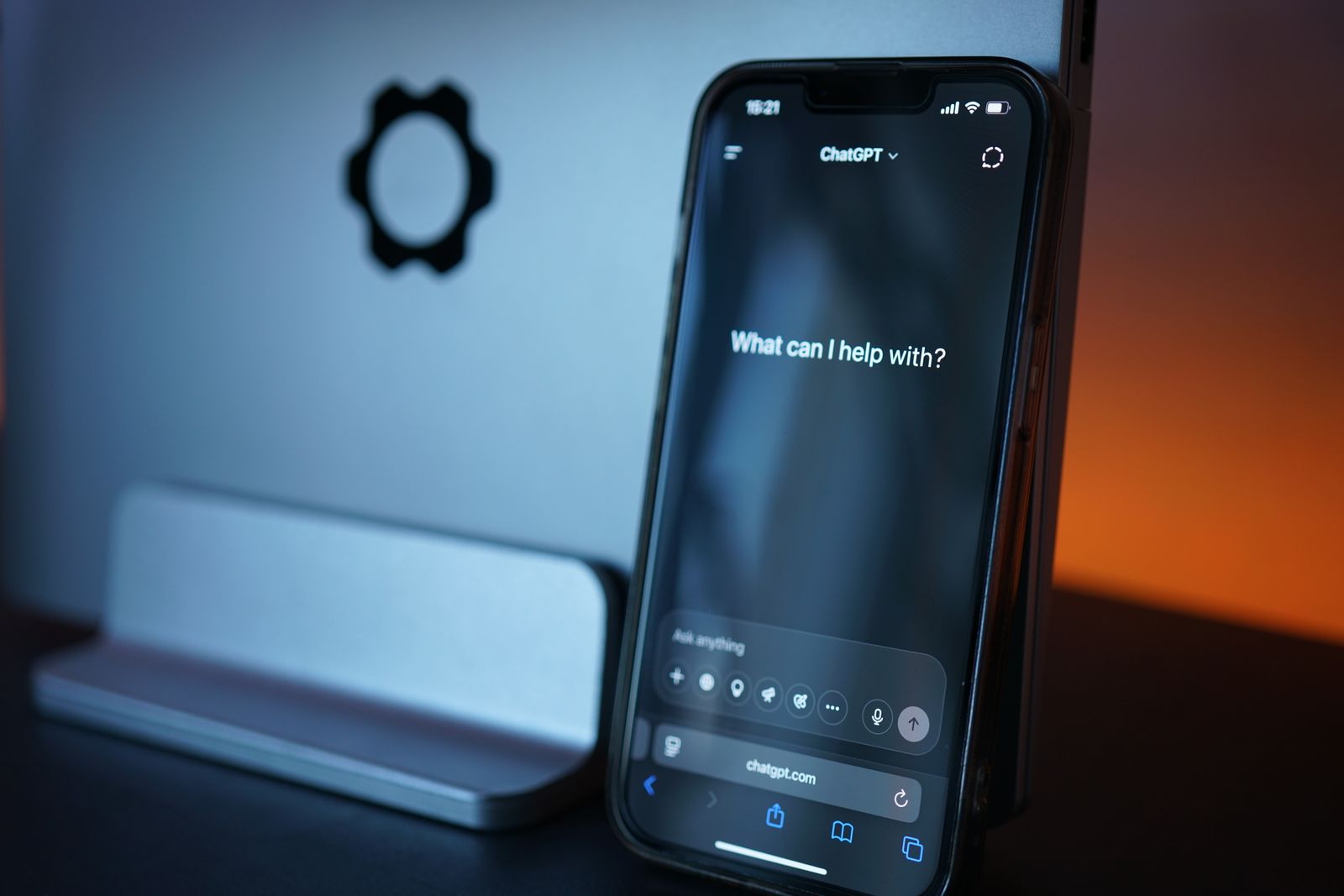 Een smartphone toont de ChatGPT-app met de openingsvraag 'What can I help with?', voor een computerscherm in een donker verlichte ruimte